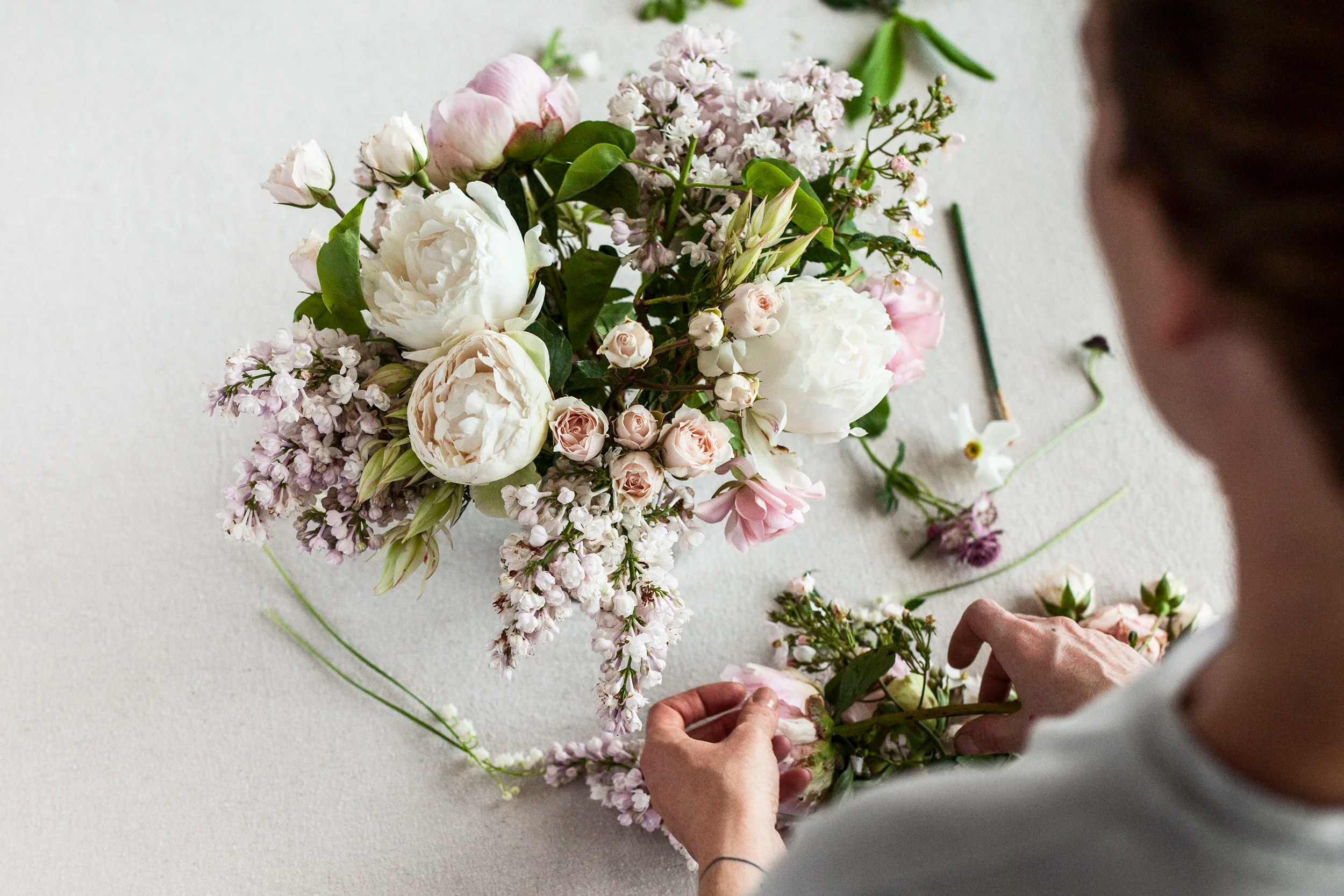 Person arranging a bouquet of light pink and white flowers on a white surface.