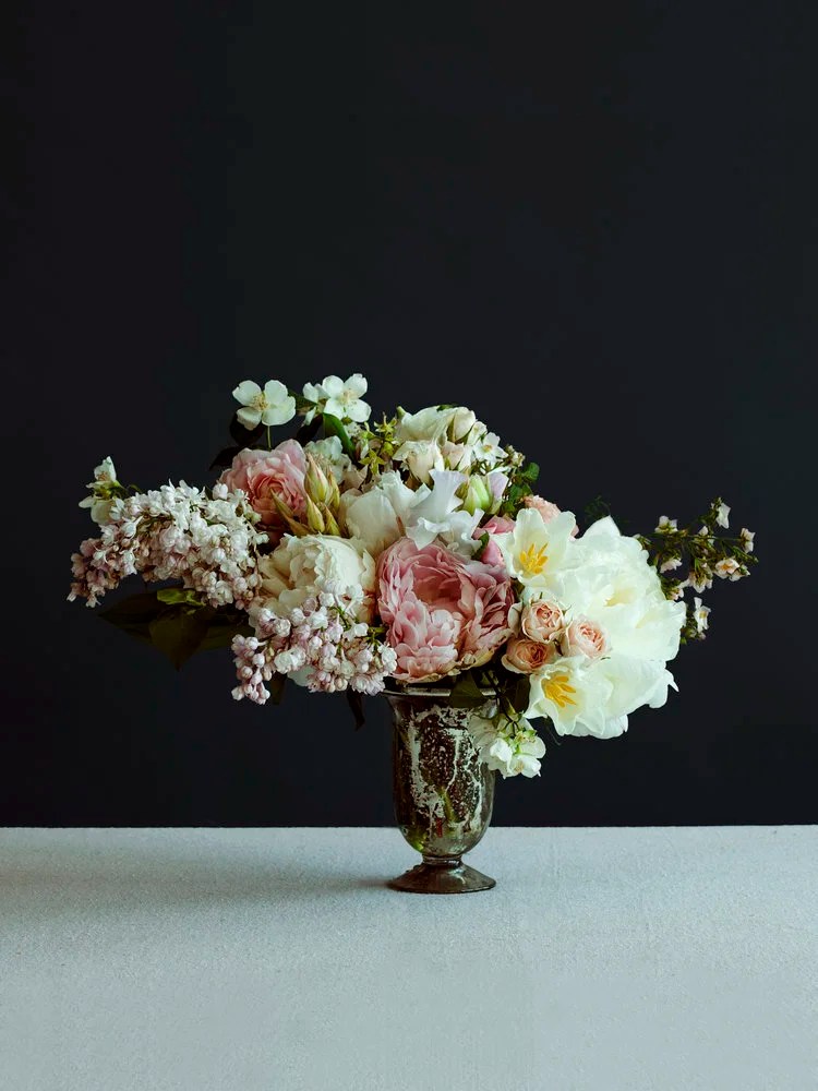 A bouquet of pink, white, and cream flowers in a dark green vase on a white surface against a dark background.